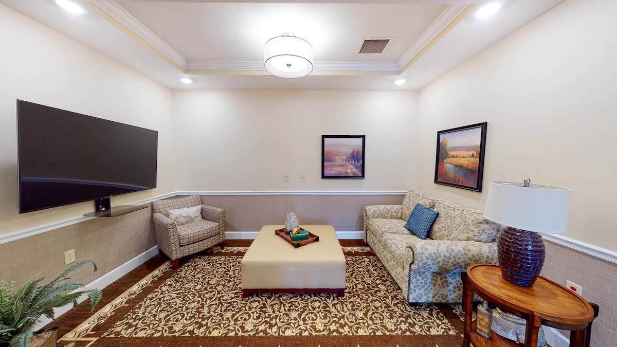 A cozy living room with a sofa, armchair, and coffee table, adorned with artwork and a patterned carpet.