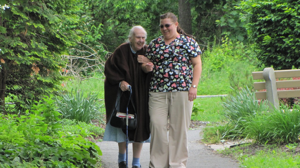 An elderly woman walking outdoors with the assistance of a caregiver.