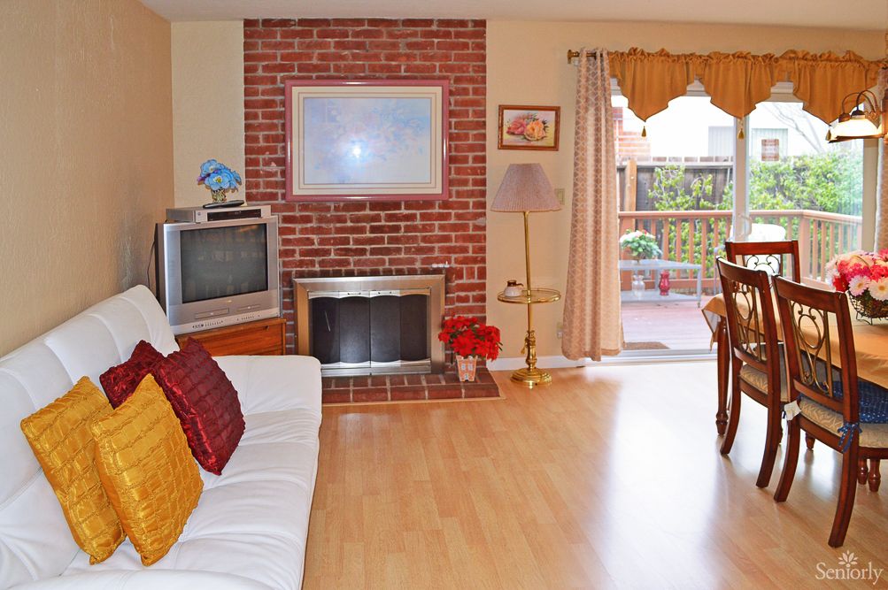 A cozy living room featuring a brick fireplace, television, and a comfortable sofa, with an adjoining dining area leading to a patio.
