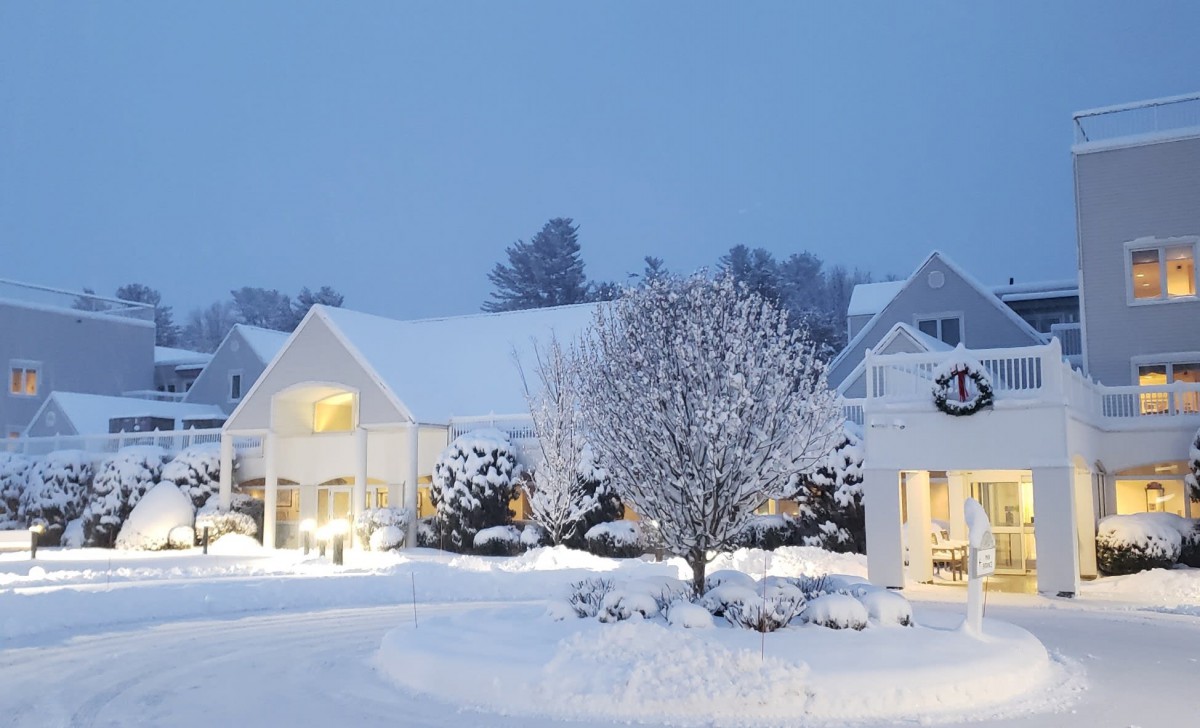A snowy evening scene with houses and trees covered in snow, illuminated by warm lights.