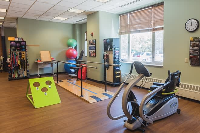 A rehabilitation therapy room equipped with an elliptical machine, exercise balls, and parallel bars.