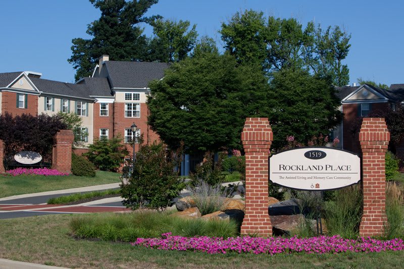 A scenic view of a brick building with a sign and well-maintained landscaping, featuring trees and colorful flowers.
