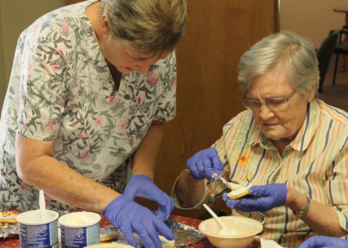 A caregiver assists an elderly woman with food preparation, both wearing gloves for hygiene.