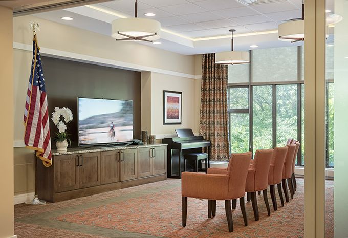 A well-decorated living room featuring a large TV, comfortable chairs, and a piano, with natural light streaming through large windows.