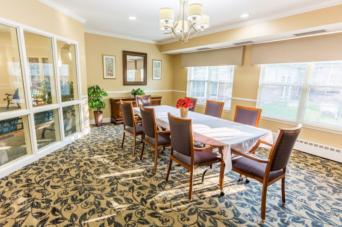 A well-lit dining room with a large table, surrounded by chairs, featuring a patterned carpet and elegant decor.