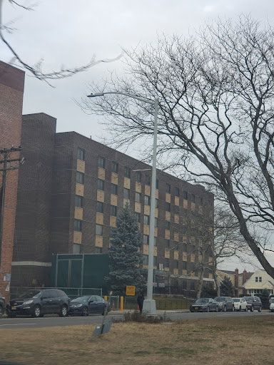 A view of a multi-story building surrounded by trees and parked cars, under a cloudy sky.