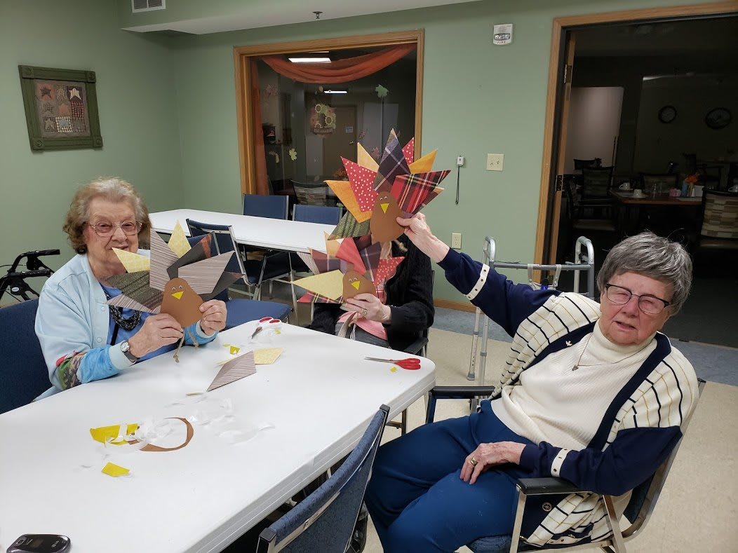 A group of seniors engaged in an indoor autumn-themed craft activity, displaying their handmade creations.