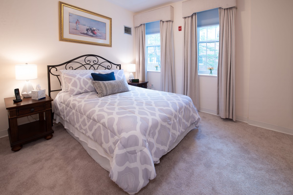 A cozy bedroom with a neatly made bed, two windows with curtains, and a bedside lamp.