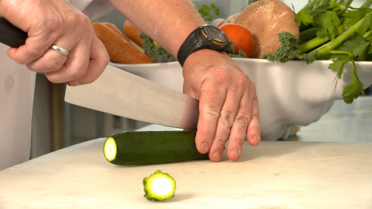 A person chopping a zucchini with a knife on a cutting board.