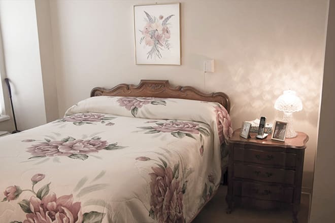 A cozy bedroom features a floral-patterned bedspread and classic wooden furniture, accompanied by a softly lit lamp on the nightstand.