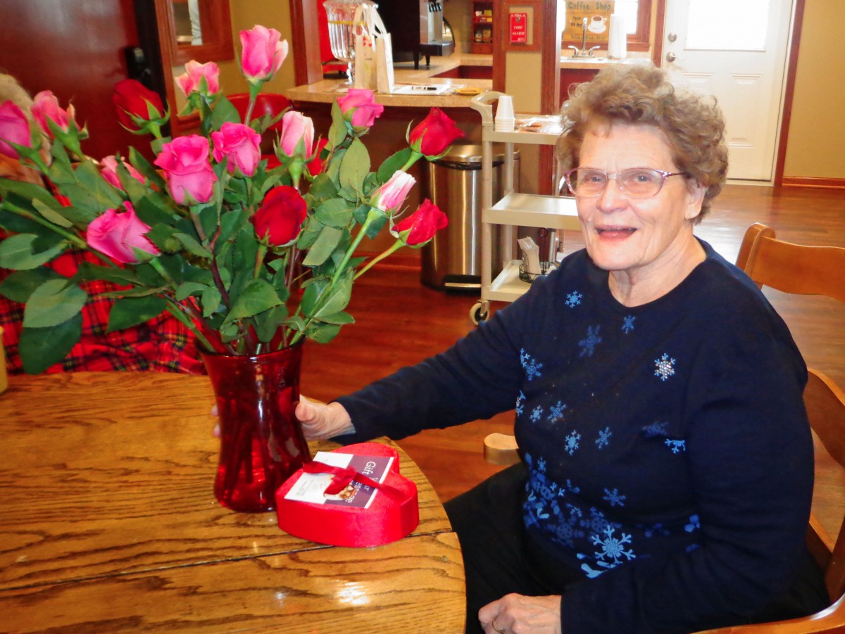 An elderly woman sitting at a table with a vase of red roses, smiling and enjoying her gift.