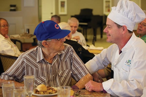 A chef engaging in a friendly conversation with an elderly man at a dining table.