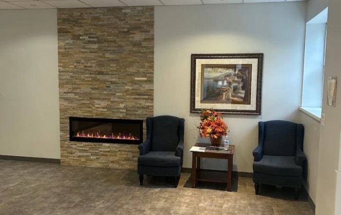 A cozy waiting room with comfortable chairs, a modern fireplace, and decorative artwork on the wall.