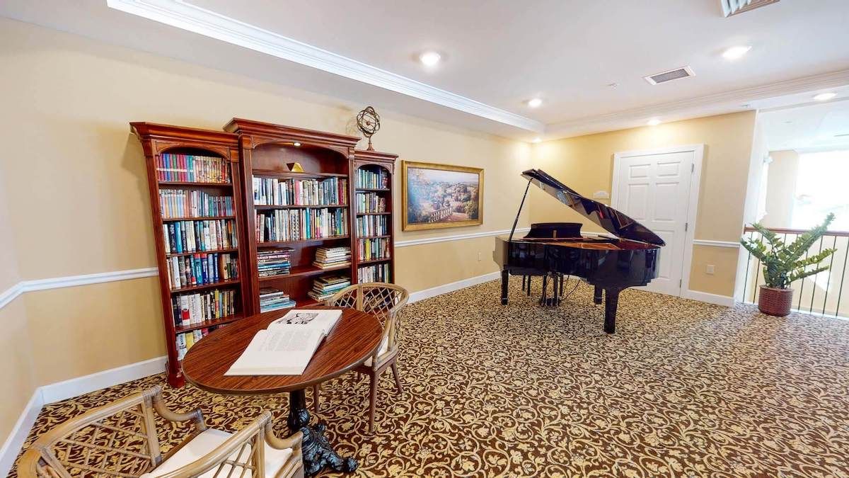 A cozy room featuring a grand piano, bookshelf filled with books, and a small table with chairs on a patterned carpet.