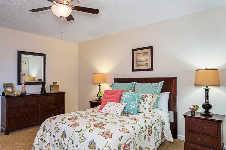 A cozy bedroom featuring a bed with colorful pillows and floral bedding, accompanied by a wooden dresser and a lamp, all under a ceiling fan.