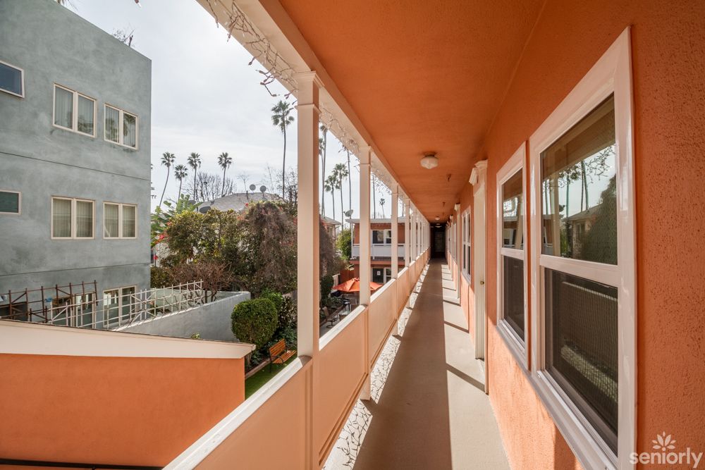 A view of an apartment building corridor with orange walls, overlooking palm trees and a residential area.