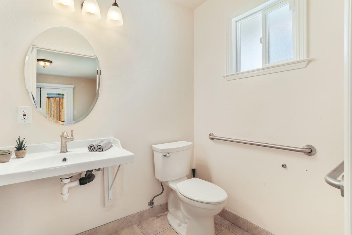 A modern, minimalist bathroom featuring a toilet, sink, mirror, and handrail next to a small window.