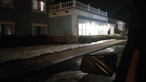 A nighttime view of a building with illuminated windows, snow along the sidewalk, and a parked car in the foreground.