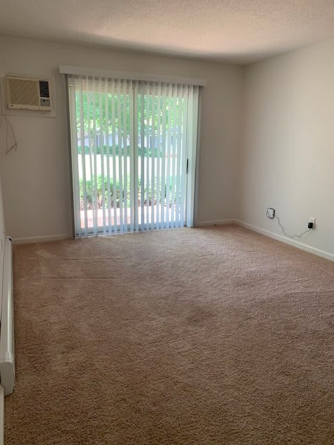 An empty room with carpet flooring, a sliding glass door with vertical blinds, and an air conditioner.