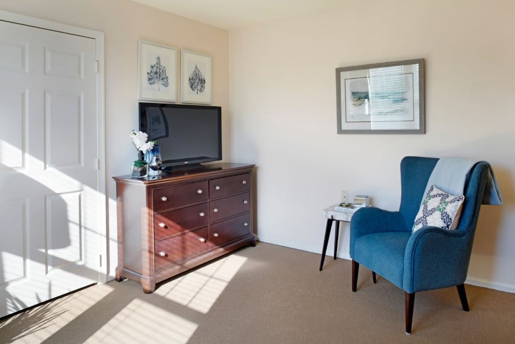 A cozy living room corner featuring a wooden dresser with a television on top, a blue armchair, and framed artwork on the walls.
