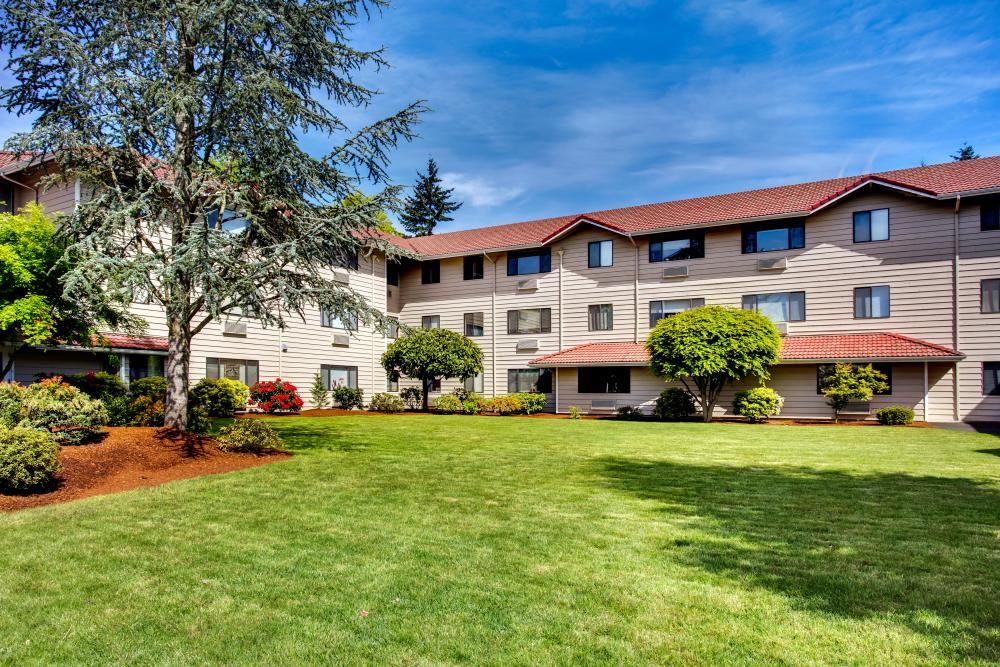 A residential apartment building with well-maintained lawns and surrounding trees under a clear blue sky.