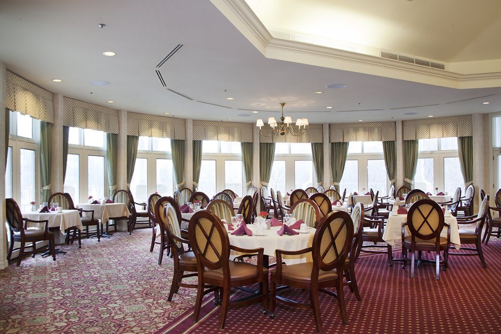 Elegant dining room with round tables and upholstered chairs, accented by large windows and a central chandelier.