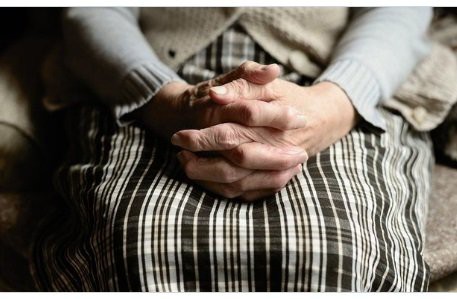 An elderly person with folded hands resting on a plaid skirt.