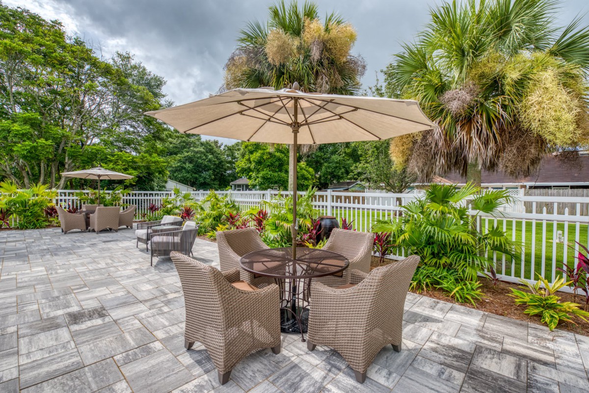 A serene patio area with wicker chairs and a table under a large umbrella, surrounded by lush greenery.