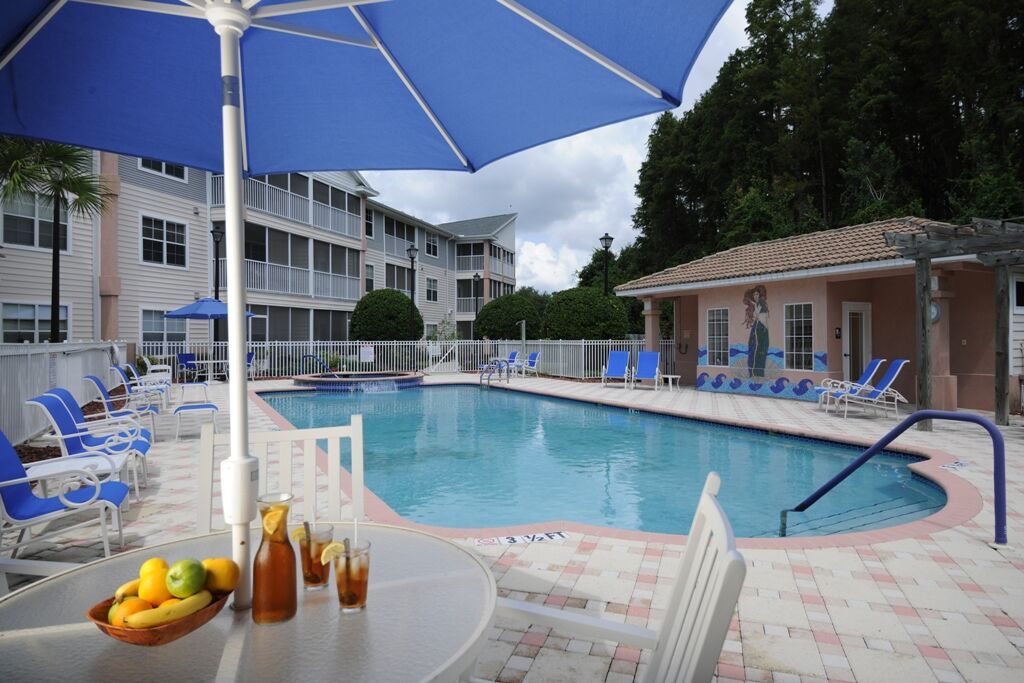 A relaxing outdoor swimming pool area with an umbrella, refreshments on a table, and surrounding resort facilities.
