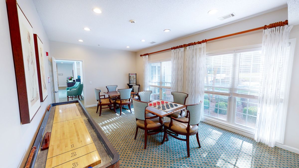 A well-lit game room featuring a shuffleboard table, seating area, and large windows with curtains.
