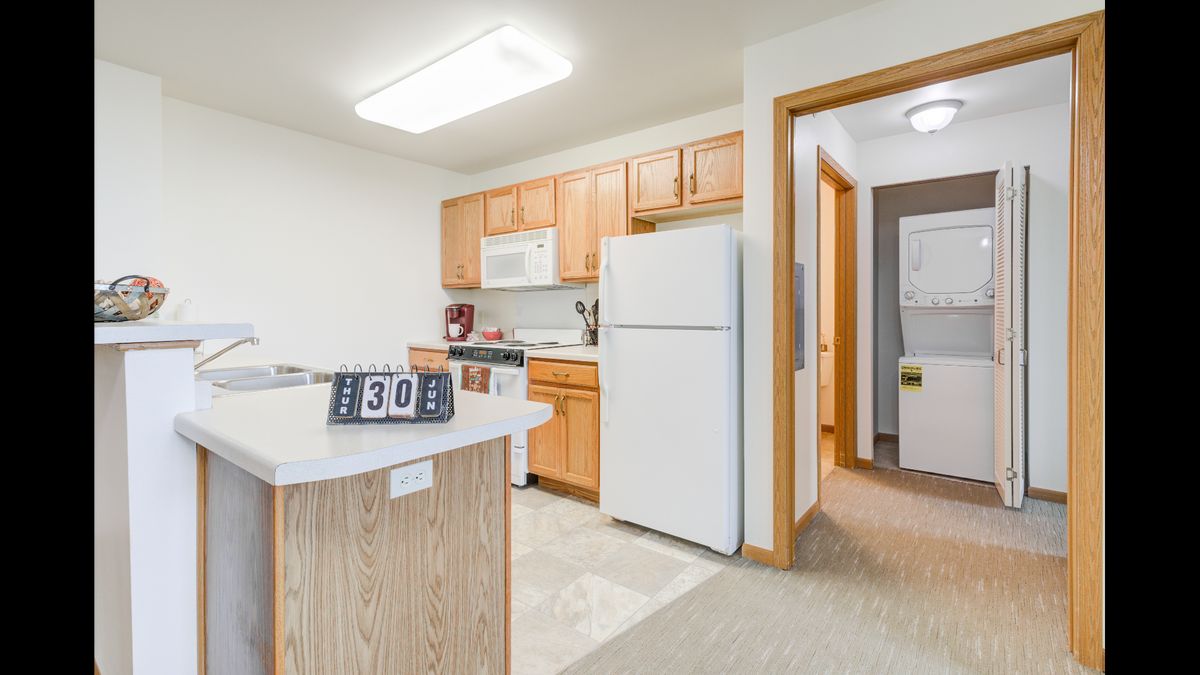 A modern apartment kitchen with wooden cabinets and white appliances.