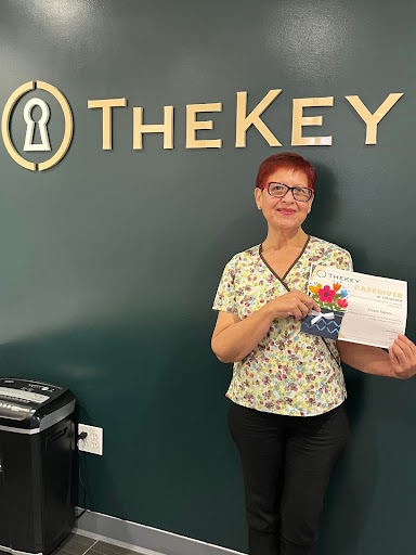 A smiling individual holding a document in front of a 'TheKey' sign in an office setting.