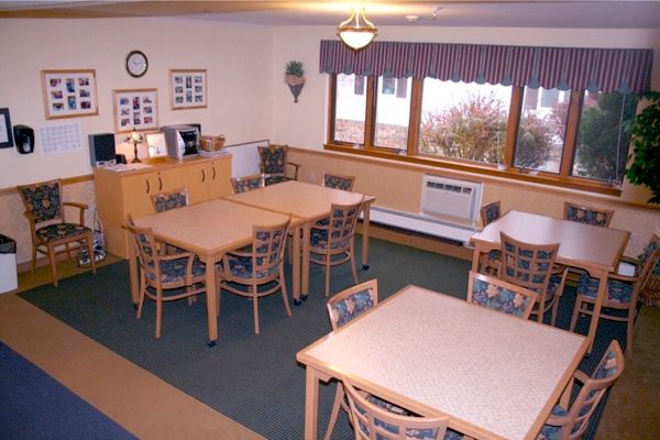 A cozy dining room with tables and chairs arranged neatly, featuring large windows and a cabinet.