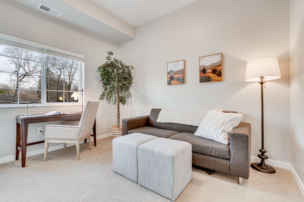A cozy living room featuring a gray sofa, two ottomans, a dining area by the window, and decorative wall art.