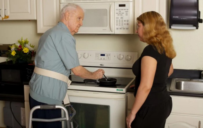 An elderly man using a walker receives assistance from a caregiver in the kitchen.