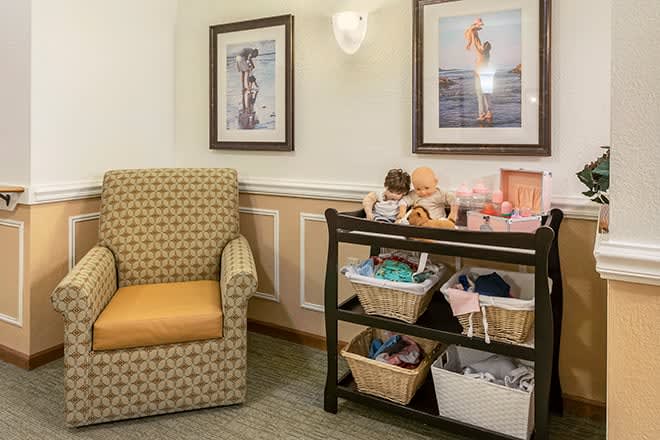 A cozy nursery corner with a comfortable chair, artwork on the wall, and a storage cart filled with baby supplies.