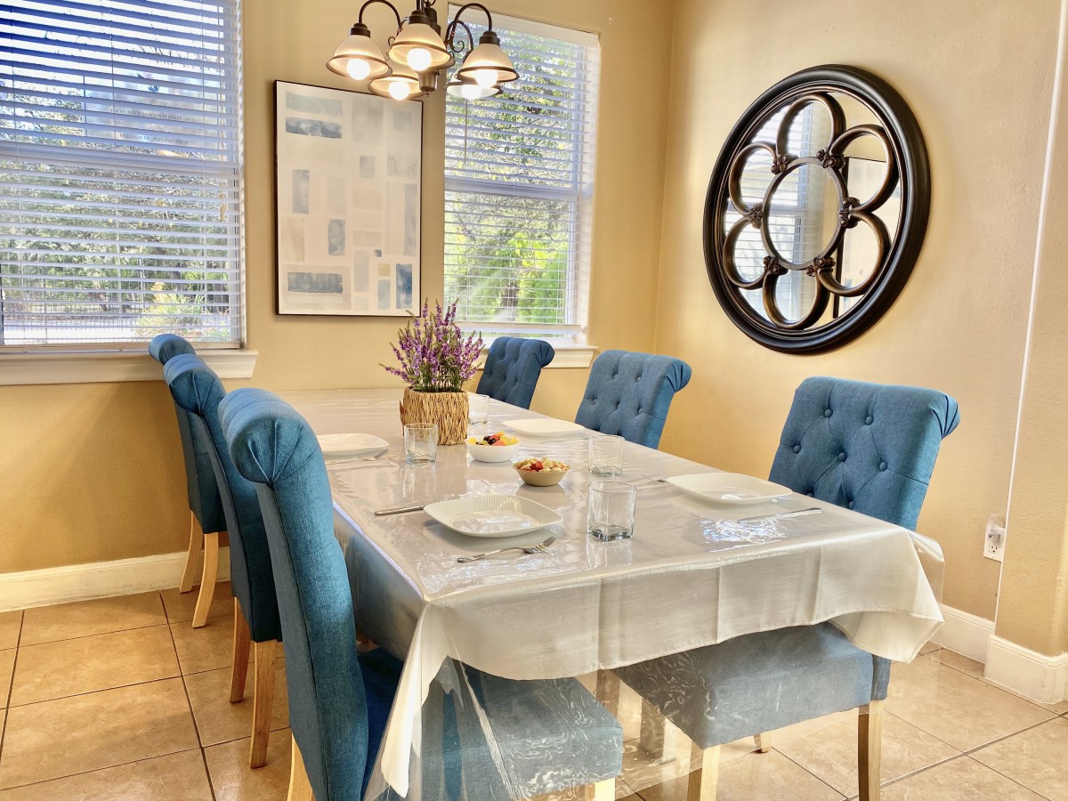A modern dining room with a neatly set table and blue upholstered chairs, featuring a decorative wall mirror and ample natural light.