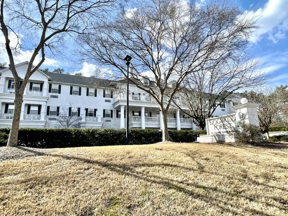 A large white building with black shutters stands behind leafless trees on a sunny winter day.