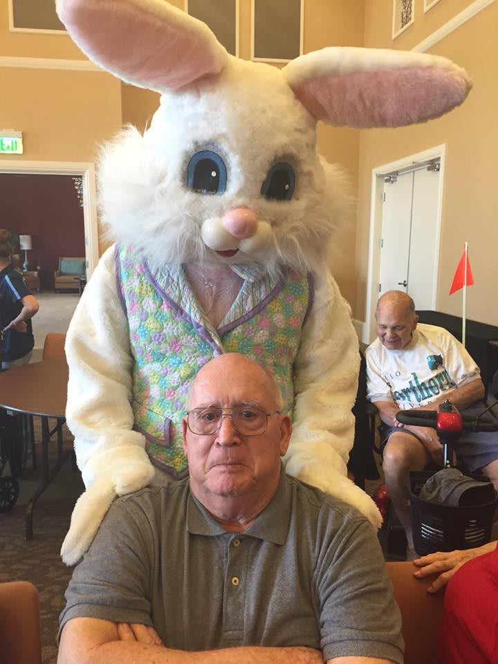 An elderly man seated with the Easter Bunny standing behind him in a room.
