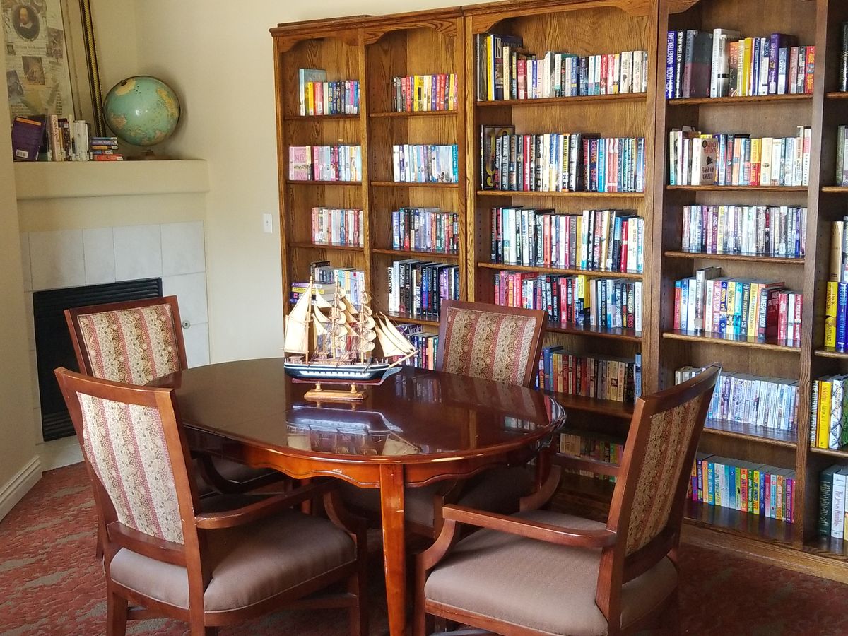 A cozy library room with wooden bookshelves filled with books, a round table surrounded by chairs, and decorative items including a globe and a model ship.