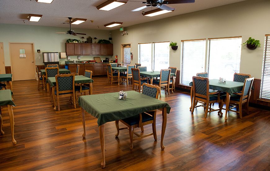 A spacious dining room with several tables and chairs, featuring a wooden floor and a kitchen area in the background.