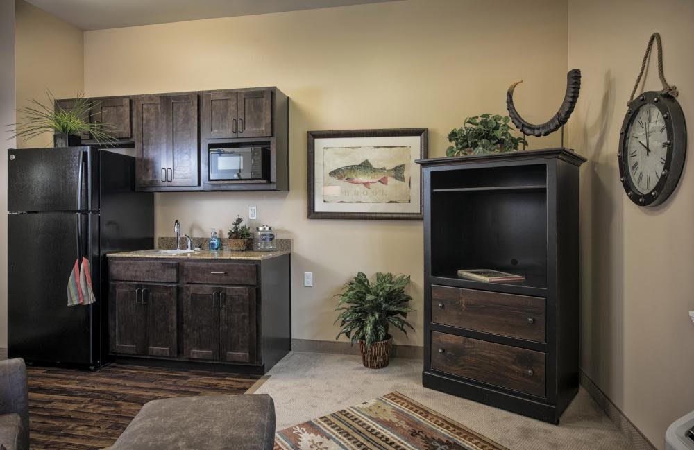 A cozy kitchenette area with dark wooden cabinets, a microwave, and rustic decor, including a fish painting and indoor plants.