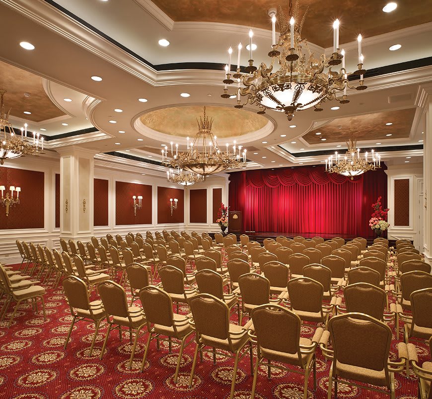 An elegant ballroom set up with rows of chairs facing a stage with red curtains, featuring ornate chandeliers and intricate ceiling design.