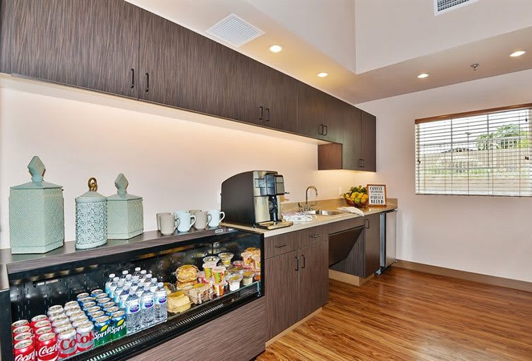 A modern kitchen space featuring a beverage station with a coffee machine, snacks, and a small refrigerator stocked with drinks.