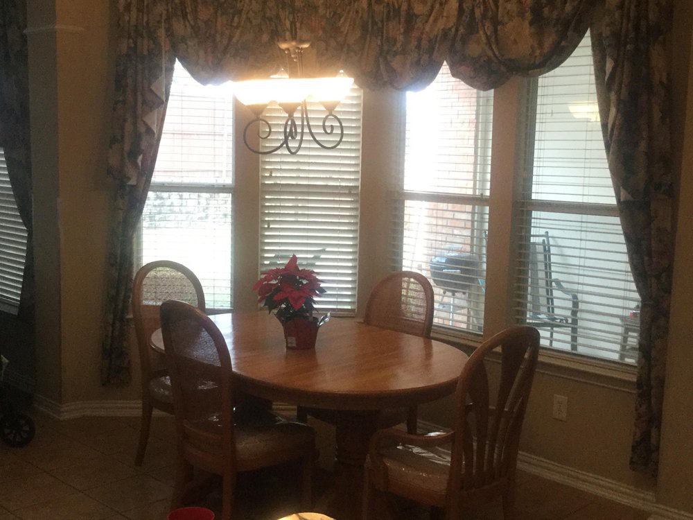 A cozy dining room with a wooden table and chairs, adorned by a chandelier and surrounded by large windows with curtains.