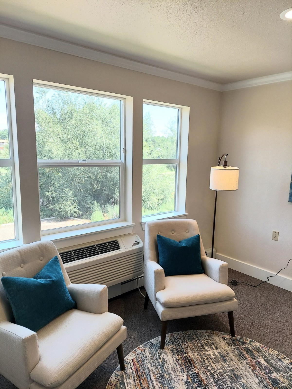 A cozy living room with chairs, a lamp, and large windows providing natural light.