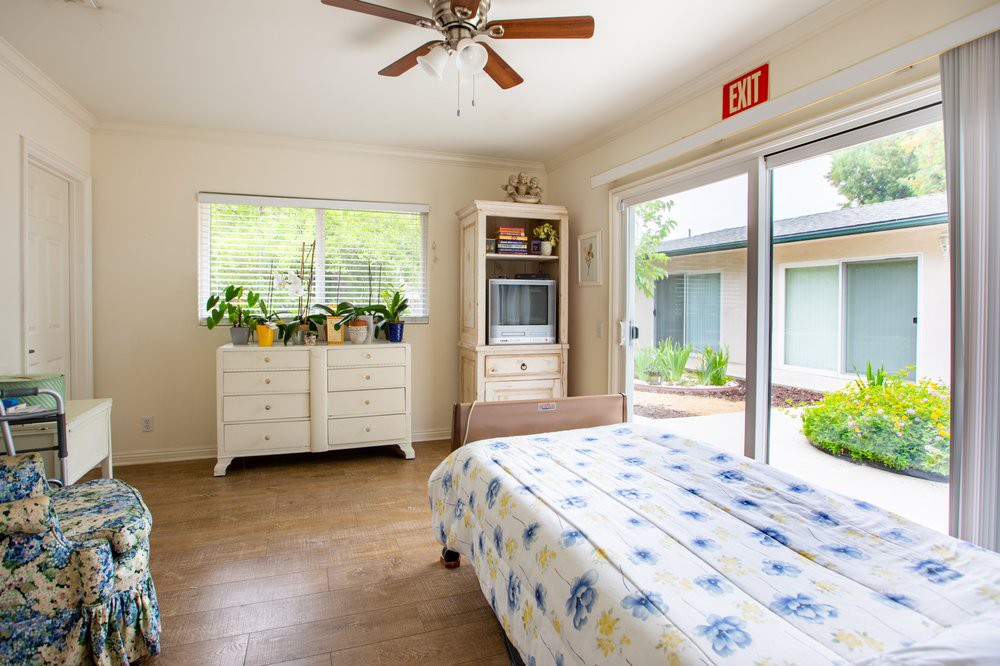 A bright bedroom with a ceiling fan, large windows, and sliding doors opening to the outside. It features a dresser with plants and floral bedding.
