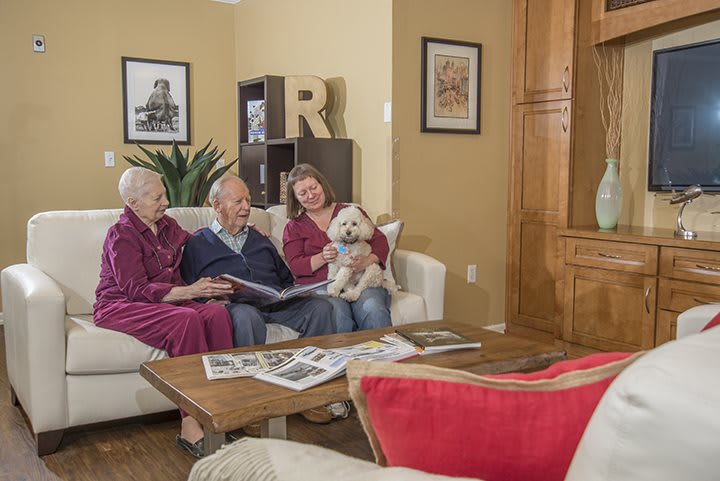 An elderly couple sits on a sofa with a younger woman holding a small dog, enjoying a cozy living room setting.