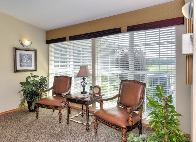 A cozy living room corner with two chairs, a small table, and a lamp by large windows with blinds.