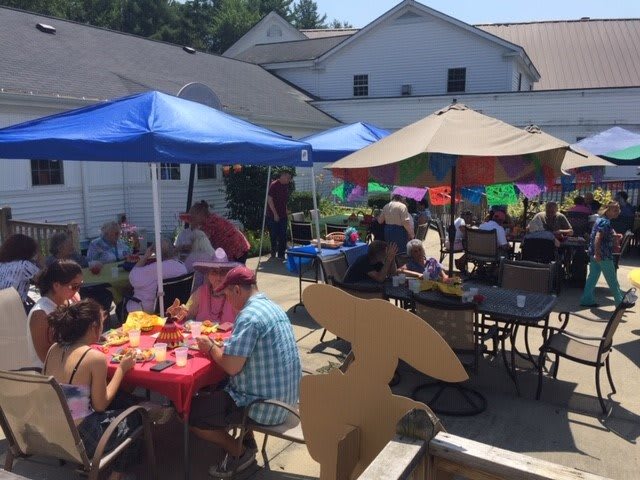 A lively outdoor gathering with people seated under umbrellas and tents, enjoying a sunny day.